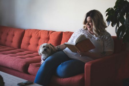 Woman sitting on red couch reaching book and petting small dog beside her